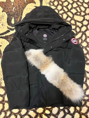 Svart dunjacka från Canada Goose - Säljer en svart dunjacka från Canada Goose med klassisk patch på ärmen. Jackan har huva med avtagbar pälskant, dragkedja och knappar framtill samt två stora fickor. Perfekt för kalla vinterdagar och riktigt snygg streetstil. Den är helt ny