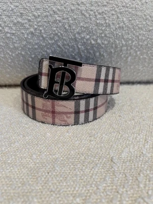  Burberry-bälte - Snyggt Burberry bälte i skinn med klassiskt rutigt mönster i beige, svart och rött på ena sidan och mörkt rutigt mönster på andra sidan. Bältet har en stor metallspänne med Burberrys ikoniska TB-logga. Perfekt accessoar för att lyfta din outfit. Man kan vända på det så att det blir 2 färger, svart och beige , kom dm för eventuella frågor 🧐 pris kan också diskuteras 💯.