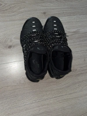 Svarta Nike sneakers med snörning - Säljer ett par svarta Nike sneakers med coola reflekterande snören och mesh-överdel. Skorna har en chunky sula med synliga luftkuddar och silverdetaljer på sidan. Perfekta för dig som gillar sportig stil och vill sticka ut med snygga detaljer.