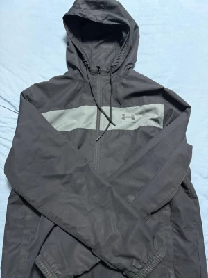 Svart vindjacka från Under Armour - Säljer en svart vindjacka från Under Armour med grått band över bröstet och logga. Jackan har huva med snörning, dragkedja framtill och långa ärmar med resår vid handlederna. Perfekt för blåsiga dagar och sportiga outfits.