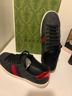 Gucci sneakers med röd/blå rand - Snygga svarta sneakers från Gucci med klassisk röd och marinblå rand på sidan. Skorna har vit platt sula, svarta skosnören och diskret GG-mönster i tyget. Baksidan har en metallicröd detalj. Tillverkade i Italien och perfekta för dig som vill sticka ut.