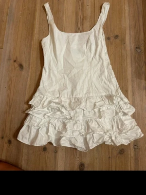 Vit volangklänning från Zara - Supersöt vit kort klänning från Zara med breda axelband och flera lager volanger nedtill. Klänningen har en dragkedja i sidan för enkel påklädning och är gjord i ett lätt bomullsmaterial. 