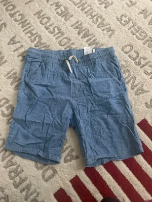 Blå shorts med snörning från H&M - Säljer ett par ljusblå shorts från H&M i storlek 164. De har en bekväm midja med vit snörning och två sidofickor. Materialet är mjukt och ser ut att vara bomull eller linne, perfekt för varma dagar. Enkla och stilrena för en chill look.