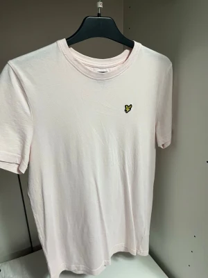 Ljusrosa t-shirt från Lyle & Scott - Säljer en ljusrosa t-shirt från Lyle & Scott Junior i 100% bomull. Klassisk passform med korta ärmar och rund hals. På bröstet sitter den ikoniska broderade gula örnen som detalj. Perfekt basic-plagg till garderoben.