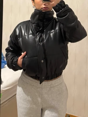 Svart pufferjacka i skinnimitation - Säljer en svart pufferjacka i skinnimitation med quiltad design. Jackan har hög krage, tryckknappar framtill och elastisk midja. Perfekt för dig som gillar en cool och trendig look. Jackan är vadderad och har långa ärmar med en mindre fläck vid fickorna.  (Se bilder ), ingeting som syns tydligt.