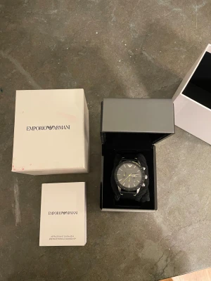 Svart Emporio Armani klocka - Säljer en stilren svart Emporio Armani klocka med läderarmband och detaljer i gult. Urtavlan har kronograf och tachymeter-funktion, samt datumvisning. Kommer med originalask och manual. Perfekt accessoar för dig som gillar snygga detaljer. Har lite spricka i glaset 