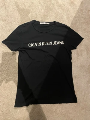 Svart t-shirt från Calvin Klein - Svart t-shirt från Calvin Klein med vit logotyptryck på bröstet. Klassisk rund hals och korta ärmar. T-shirten har en clean och stilren look, perfekt för dig som gillar enkla och snygga plagg. Materialet känns mjukt och skönt mot huden.