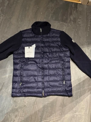 Moncler cardigan - Snygg mörkblå dunväst från Moncler med quiltad framsida och stickade ärmar. Västen har två dragkedjefickor, hög krage och hel dragkedja framtill. Materialet är polyester och dun, med Moncler-logga på ärmen och coolt serietryck på insidan.