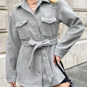 Grå overshirt med bälte - Säljer en grå overshirt med knappar framtill och två stora bröstfickor. Jackan har ett snyggt bälte i midjan som knyts och ger en cool siluett. Materialet är mjukt och känns skönt mot huden. Perfekt för lager-på-lager och enkel att styla. (Färgen är som på bild 1, ljusfel på min egna bild 2.)