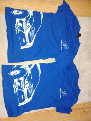 Blå Petter Solberg t-shirt med biltryck - Säljer en blå t-shirt med coolt biltryck och texten 'Pushing the limits' på framsidan. Baksidan har ett stort vitt tryck med Petter Solberg-logga. T-shirten har rund hals och korta ärmar, perfekt för dig som gillar motorsport och rally.