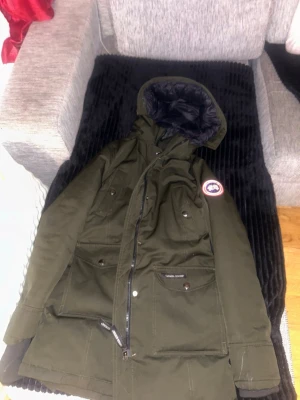 Mörkgrön dunparkas från Canada Goose XS - Säljer en mörkgrön parkajacka från Canada Goose i storlek XS. Jackan har huva, flera fickor med knappar och dragkedja framtill. Insidan är quiltad och fodrad med dun för extra värme. Perfekt för kalla dagar och har klassisk Canada Goose-logga på ärmen.