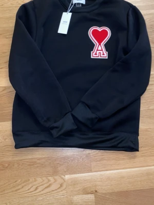Svart AMI Paris sweatshirt med hjärtlogga - Stilren svart sweatshirt från AMI Paris med den ikoniska röda hjärtloggan med “A” på bröstet. Klassisk passform med rund hals och mjukt, bekvämt material. Ett tidlöst och exklusivt plagg som passar perfekt till både vardag och mer uppklädda outfits. Tag kvar – ger ett fräscht och lyxigt intryck.