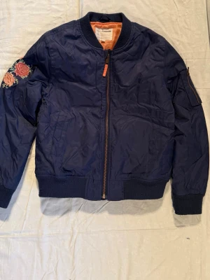 Blå bomberjacka med broderade blommor - Snygg mörkblå bomberjacka från KappAhl med broderade blommor i rosa och grönt på ryggen och ena ärmen. Jackan har dragkedja framtill, ribbade muddar och en cool orange insida. Perfekt för dig som vill sticka ut med detaljer!