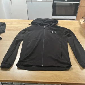 Svart vindjacka från Under Armour - Svart vindjacka från Under Armour med huva och hel dragkedja framtill. Jackan har elastiska muddar vid ärmsluten och en diskret logga på bröstet. Perfekt för dig som gillar sportig stil och vill ha något lätt och smidigt till vardags.