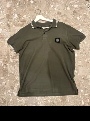 Olivgrön piké från Stone Island - Säljer en olivgrön pikétröja från Stone Island med klassisk krage och vita ränder på krage och ärmslut. Tröjan har korta ärmar, två knappar framtill och en broderad logotyp på bröstet. Materialet är mjuk bomull som känns skön mot huden.