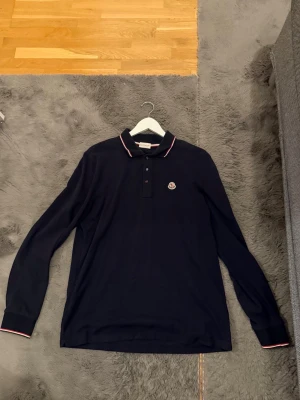 Svart långärmad polotopp från Moncler - Säljer en svart långärmad polotopp från Moncler med klassisk krage och knappar framtill. Toppens krage och ärmslut har tunna ränder i vitt och rött. Moncler-logga broderad på bröstet. Materialet är mjukt och känns lyxigt. OBS storlek är XL men sitter bra på dig som bär L 
