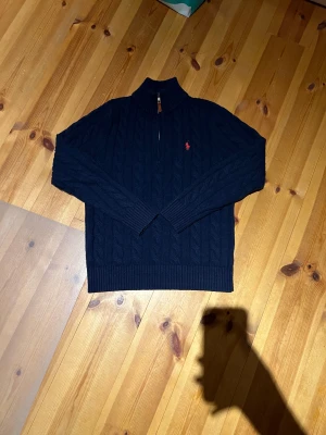 Mörkblå stickad polotröja Polo Ralph Lauren - Snygg mörkblå stickad polotröja från Polo Ralph Lauren med klassiskt kabelmönster och röd broderad logga på bröstet. Tröjan har half zip med läderdetalj, ribbade muddar och hög krage. Perfekt för höst och vinter, riktigt chill och stilren.