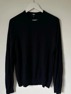 Mörkblå stickad tröja från Uniqlo - En stilren mörkblå stickad tröja från Uniqlo i storlek M. Tröjan har rund hals och långa ärmar, perfekt för lager-på-lager under kyliga dagar. Materialet är 100% ull vilket gör den både mjuk och varm. Enkel design utan mönster eller detaljer. 5L14