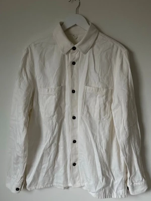 Vit överskjorta i denim från ARKET - Säljer en stilren vit overshirt från ARKET i 100% ekologisk bomullsdenim. Skjortan har klassisk krage, svarta knappar framtill och två stora bröstfickor. Passar dig som gillar enkel och clean look med långärmad design. 6Q9