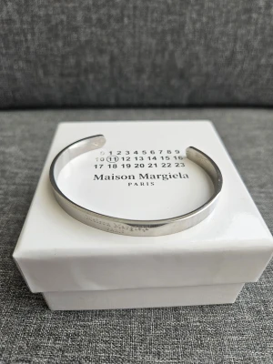 Maison Margiela silverfärgad armband - Snyggt och stilrent armband från Maison Margiela i silverfärgat 925 sterling silver. Designen är minimalistisk med en graverad logga och öppen form som gör det enkelt att ta på och av. Perfekt accessoar för dig som gillar exklusiva detaljer.