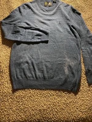 Blå stickad tröja från Lyle & Scott - Snygg blå stickad tröja från Lyle & Scott med rund hals och diskret broderad logga på bröstet. Tröjan har ribbade muddar vid ärmslut och nederkant, perfekt för lager-på-lager under kyliga dagar. Passar dig som gillar klassisk stil med modern touch. Litet hål i bröstet DÄRAV PRISET! Fler frågor? Bara o hojta till ! 