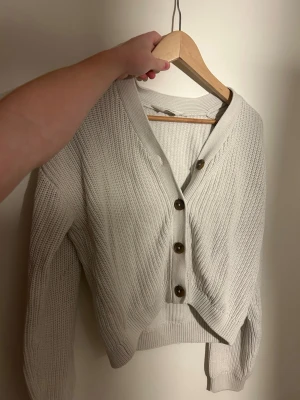 Beige stickad kofta från H&M - Säljer en beige stickad kofta från H&M i storlek 158/164. Koftan har grov ribbstickning, v-ringad hals och fyra bruna knappar framtill. Materialet är 60% bomull och 40% akryl, vilket gör den mjuk och skön. Perfekt att slänga på sig när det är lite kyligt ute.