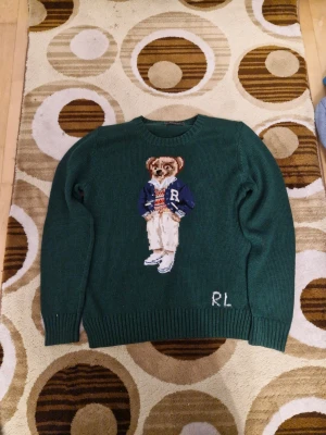 Grön stickad tröja Polo Ralph Lauren - Snygg mörkgrön stickad tröja från Polo Ralph Lauren med ikonisk björn på bröstet och RL-broderi. Rund hals och ribbade muddar. Tillverkad i Italien med merinoull, polyester, akryl och nylon. Perfekt för dig som vill ha en unik och trendig look.