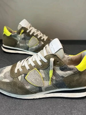 Philippe Model sneakers med camomönster - Säljer ett par Philippe Model sneakers med grått och grönt camomönster, detaljer i neon-gult och vita snören. Skorna har en rund tå, platt sula och är tillverkade i en mix av mocka och textil. Märkets logga syns på sidan och hälen har en cool neon-detalj.