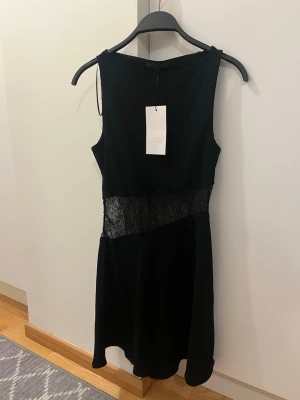 Svart klänning med spetsdetalj från Zara - Snygg svart kortklänning från Zara med ärmlös design och en transparent spetsdetalj i midjan som ger en extra touch. Klänningen har en enkel, klassisk form och passar perfekt till dig som vill ha något stilrent men ändå lite edgy.