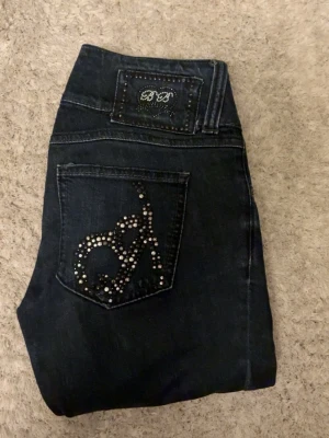 Mörkblå jeans med nitar på fickor - Säljer ett par mörkblå jeans med lite bootcut och snygga nitar i mönster på bakfickorna. Jeansen har dubbla knappar i midjan och dekorativ patch bak. Perfekta för dig som gillar detaljer och vill sticka ut lite extra.