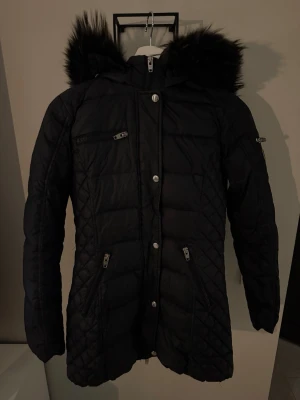 Svart Rockandblue dunjacka - Säljer en svart lång dunjacka från Rockandblue med stor huva och fluffig päls. Jackan har quiltade detaljer, flera dragkedjeförsedda fickor och Rockandblue-logga på ärmen.