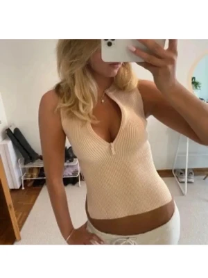 Beige ribbad ärmlös topp med dragkedja - Säljer en beige ribbad topp utan ärmar med en snygg dragkedja framtill som går att justera för djupare urringning. Toppen är croppad och har en tight passform, perfekt till jeans eller kjol. Superenkel att styla för en trendig look.