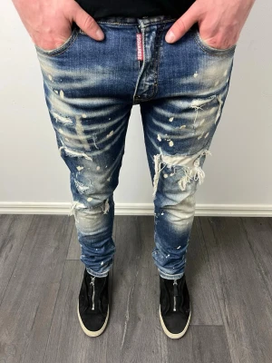 Dsquared2 blå distressed jeans - Säljer ett par blå jeans från Dsquared2 med coolt distressed utseende, slitningar och färgstänk. Jeansen har normal passform, klassisk femficksdesign och ikonisk Dsquared2-tag vid fickan. Materialet är robust jeans med snygg tvätt och detaljerade slitningar.  Hör av er kring minsta funderingar!