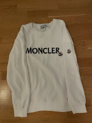 Vit sweatshirt från Moncler - Vit sweatshirt från Moncler i bomull med stor svart Moncler-logga på bröstet och två broderade logopatchar på ena ärmen. Klassisk rund halsringning och ribbade muddar vid ärmslut och nederkant. Enkel och clean design som passar till allt.