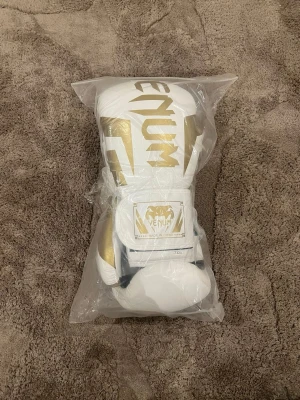 Venum vita och guld boxningshandskar - Snygga boxningshandskar från Venum i vitt med guldiga detaljer och logga. Handskarna har kardborreknäppning och är tillverkade i syntetmaterial. Perfekta för dig som vill sticka ut på träningen. Inte de bästa kvalite men fortfarande ok därför jag säljer de för eyt billigt pris