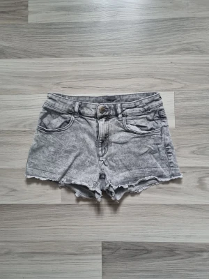 Grå denimshorts från H&M - Säljer ett par gråa denimshorts från H&M i storlek 146. De har råa, fransiga kanter och klassisk femficksdesign med knapp och dragkedja framtill. Perfekta för sommardagar och har en snygg tvättad look.