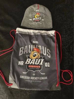 Ingen väst – SHL Bauhaus mössa & gympåse - Det här är ett set med en grå mössa och en gympåse från SHL Bauhaus Hockey Team. Mössan är i mjuk bomull med tryck och röd söm, och påsen är i grått meshmaterial med rött snöre och stort tryck framtill. Perfekt för hockeyfans!