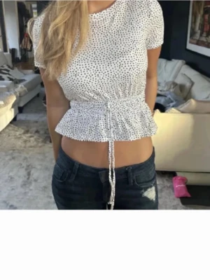 Vit prickig topp med snörning - Supersöt vit topp med små svarta prickar och snörning i midjan som ger en peplum-effekt. Kortärmad och croppad modell som passar perfekt till jeans eller kjol. Tunn och luftig, perfekt för varma dagar.