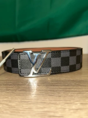 Rutig Iv bälte - Säljer ett snyggt bälte med klassiskt rutigt mönster i svart och grått. Bältet har en stor silverfärgad logga som spänne och är tillverkat i skinn med brunt insida. Perfekt accessoar för att lyfta din outfit. 