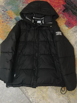Svart pufferjacka från Burberry - Säljer en svart pufferjacka från Burberry med avtagbara ärmar så du kan bära den som väst. Jackan har huva, dragkedja och knappar framtill samt två fickor med dragkedja. Klassisk Burberry-rutig detalj på insidan av huvan och logga på ärmen.