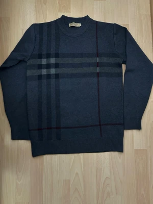 Mörkblå stickad Burberry tröja - Snygg mörkblå tröja från Burberry med klassiskt rutmönster i grått och vin sopprött. Tröjan har rund halsringning och långa ärmar. Perfekt för dig som vill ha en stilren och tidlös look med exklusiv känsla.säljer den för den är lite för stor för mig!