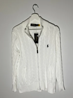 Vit kabelstickad kofta från Polo Ralph Lauren - Säljer en vit kabelstickad kofta från Polo Ralph Lauren. I storlek M. Rekommenderad längd är 175-85