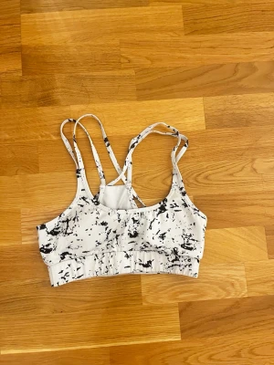 Vit och svart träningstopp med mönster - Säljer en sportig väst i vitt med svart abstrakt mönster. Västen har tunna, korsade axelband och bred resår under bysten för extra stöd. Perfekt för träning och har en modern look med sitt unika mönster. Den är helt oanvänd då jag beställde fel storlek 