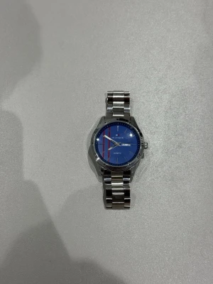 Blå Tommy Hilfiger klocka i stål - Snygg klocka från Tommy Hilfiger med blå urtavla och röda detaljer. Armbandet är i blankt rostfritt stål och har ett klassiskt spänne med TOMMY ingraverat. Urtavlan har dag- och datumvisning samt diskreta Hilfiger-loggan.