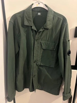 Grön overshirt från C.P. Company - Säljer en grön overshirt från C.P. Company med klassisk krage, stora bröstfickor och knappstängning. Ärmen har den ikoniska linsen som ger jackan en unik touch. Perfekt lager-på-lager-plagg för dig som gillar streetwear. Billigare vid snabbaffär, köpt på NK kvitto finns