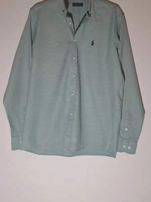 Grön skjorta från Polo Ralph Lauren M - Klassisk grön skjorta från Polo Ralph Lauren med broderad marinblå logga på bröstet. Skjortan har lång ärm, knappar framtill och på manschetterna. Tillverkad i mjuk bomull och har en avslappnad passform.