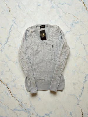 Ljusblå kabelstickad tröja Polo Ralph Lauren - Säljer en ljusblå kabelstickad tröja från Polo Ralph Lauren med klassisk v-ringning och broderad logga på bröstet. Tröjan har lång ärm och ribbade muddar, perfekt för dig som gillar stilrena och tidlösa plagg.