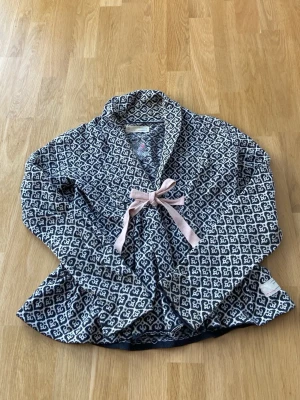 Mönstrad kofta med rosett från Odd Molly - Supersöt kofta från Odd Molly i grå och vitt med ett grafiskt mönster och en ljusrosa rosett framtill. Koftan har lång ärm och är gjord i mjuk bomull, 