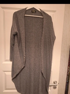 Grå kofta från Ester Elenora - Mysig och lång grå kofta från Ester Elenora. Koftan har öppen front med dekorativa nitar längs kanterna och är tillverkad i ett mjukt stickat material. Perfekt för lager på lager och passar till både jeans och klänning.