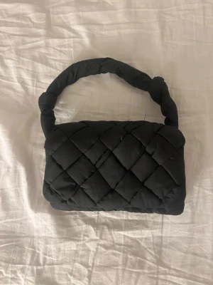 Svart quiltad handväska Carin Wester - Säljer en svart quiltad handväska från Carin Wester med vadderat material och flätat handtag. Väskan har två stora fack och en innerficka med dragkedja. Perfekt storlek för det viktigaste och riktigt snygg design.